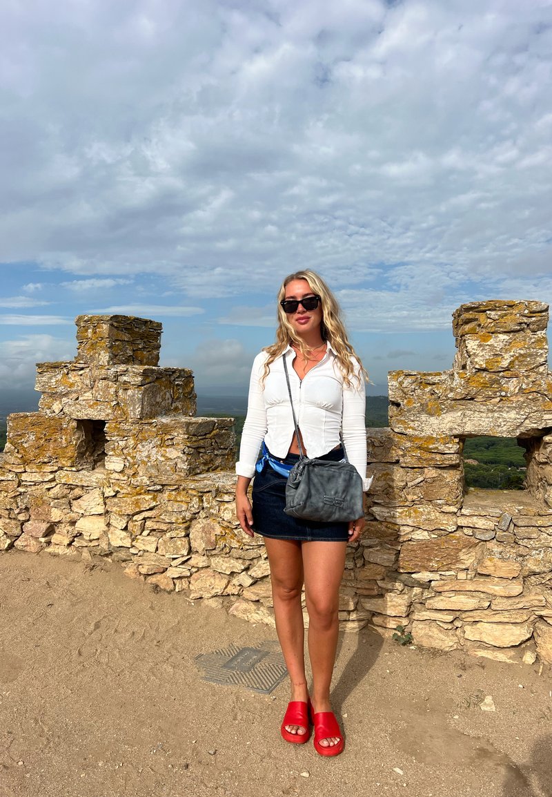 Una donna si trova vicino a un muro di pietra, indossando un top bianco con zip, una gonna di jeans blu, sandali rossi e portando una borsa a tracolla grigia.
