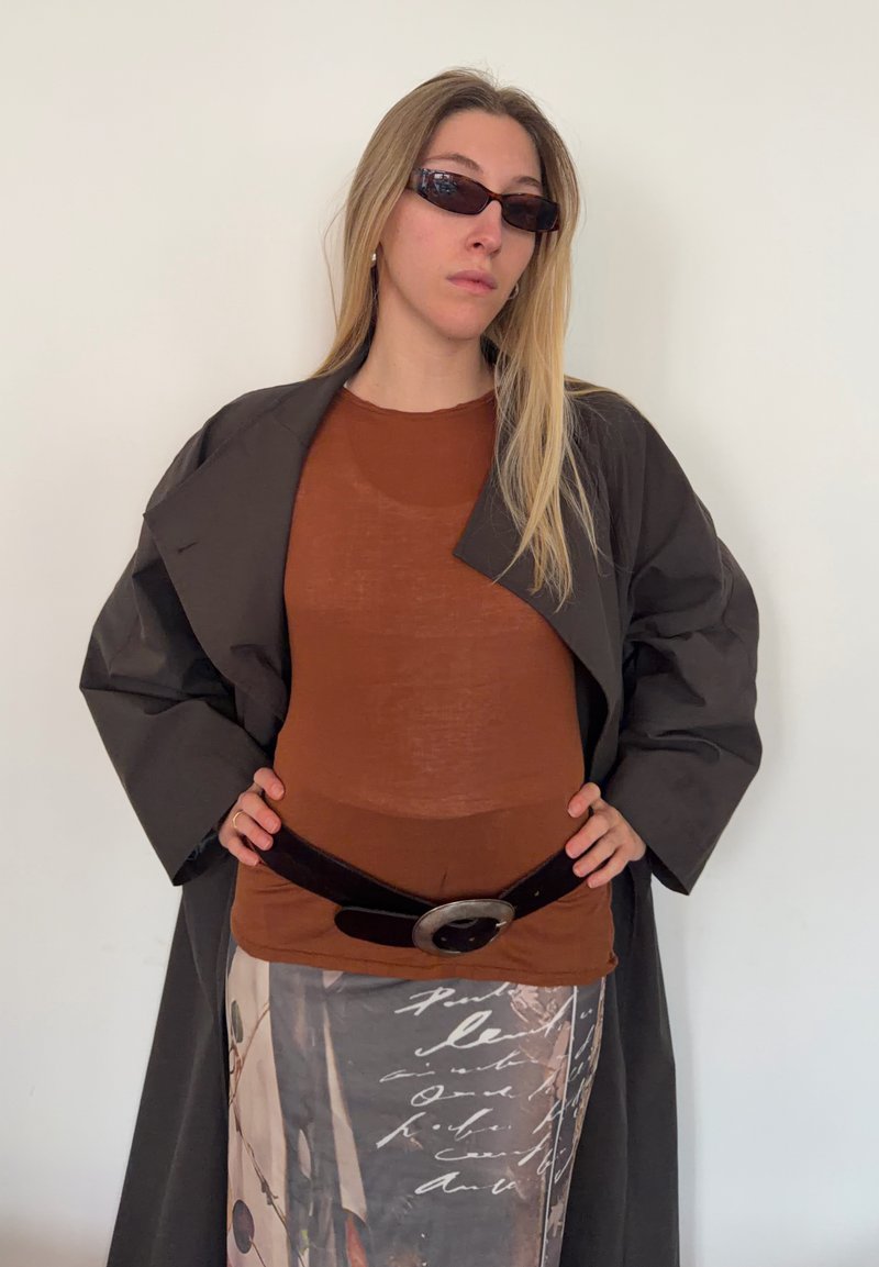 Haut marron ajusté, manteau sombre oversize, jupe à motifs aux tons terreux avec texte, large ceinture noire avec boucle argentée, lunettes de soleil rectangulaires.