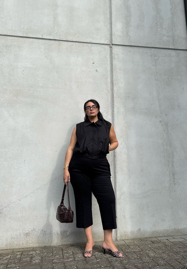 Mujer con top negro sin mangas y pantalones cortos está apoyada contra una pared de hormigón sosteniendo un bolso oscuro, lleva gafas y tacones con estampado de leopardo.