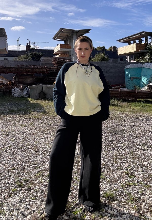 Une personne portant un haut crème et noir et un pantalon noir ample se tient dans une cour de chantier recouverte de gravier, avec des matériaux empilés et des toilettes portables.