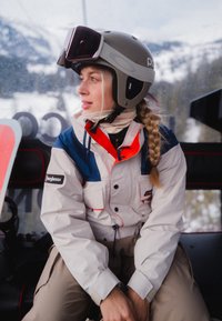 Femme aux cheveux tressés portant un casque de ski et une veste, assise à l'intérieur d'une cabine de télésiège avec une montagne enneigée visible à travers la fenêtre.