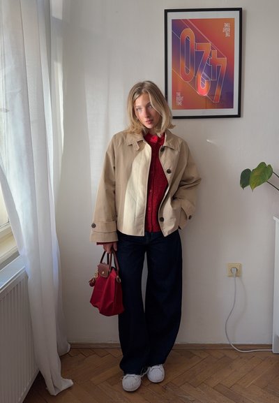 Mujer con pelo rubio vestida con chaqueta beige, suéter rojo y pantalones negros, sosteniendo un bolso rojo, de pie en una habitación con un cartel colorido enmarcado.