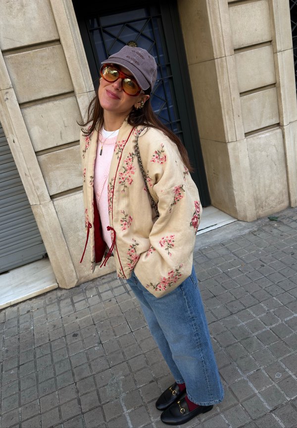 Woman wearing a floral beige jacket, pink jumper, blue jeans, grey cap, orange sunglasses, and black loafers standing on a city pavement.