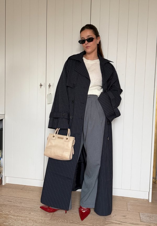 Lang zwarte trenchcoat met pinstripe, brede revers, crèmekleurige trui, grijze geplooide broek en rode pumps met puntneus, met een beige handtas in de hand.
