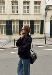 Čierny pletený sveter s vysokým golierom, v páre so širokými džínsami s rovnými nohavicami; čierna kabelka so zlatými kovaniami spočíva na ramene.