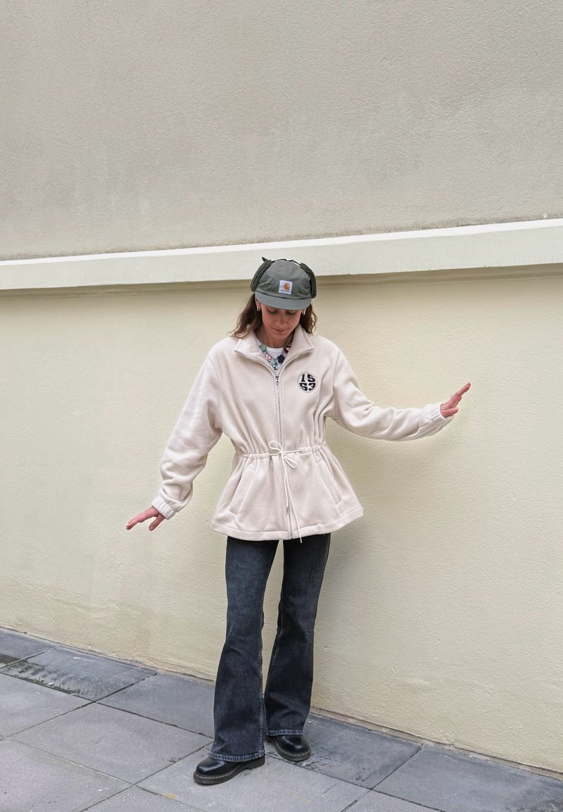 Veste en polaire crème avec taille cintrée, fermeture éclair et détail logo. Associée à un jean évasé foncé et des bottines noires. Chapeau vert avec rabats pour les oreilles.