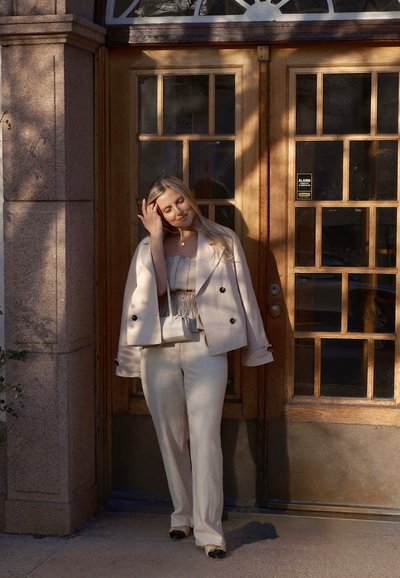 Blazer beige a medida con botones negros, una blusa de tela ligera y pantalones a juego. Enmarcado por puertas dobles de madera con un diseño de cristal.