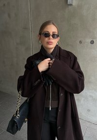 Σκούρο καφέ oversized παλτό, εφαρμοστό καφέ φούτερ με φερμουάρ, μαύρα γάντια, χρυσή τσάντα, μαύρα γυαλιά ηλίου και υφασμάτινο φόντο.