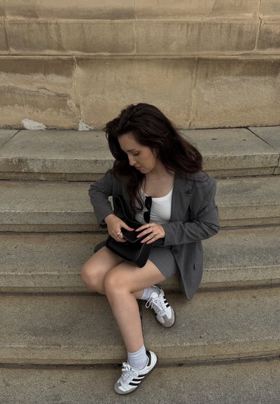 Joven mujer con blazer gris y falda sentada en escalones de piedra, abriendo un bolso negro, vistiendo zapatillas blancas y calcetines blancos.