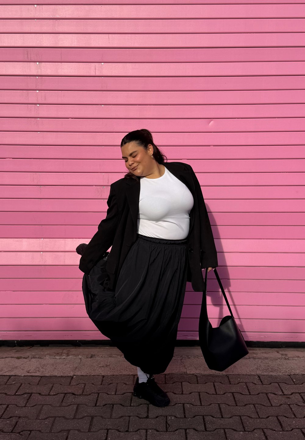 Blazer preto sobre uma t-shirt branca, saia preta fluída e mala preta contra um fundo rosa com linhas horizontais.