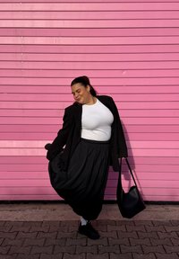 Blazer preto sobre uma t-shirt branca, saia preta fluída e mala preta contra um fundo rosa com linhas horizontais.