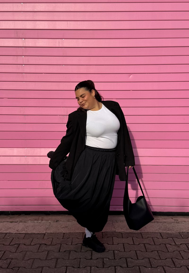Schwarzer Blazer über einem weißen T-Shirt, fließender schwarzer Rock und schwarze Handtasche vor einem pinkfarbenen Hintergrund mit horizontalen Linien.