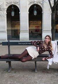 Femme portant un haut marron à pois blancs, une jupe beige et des collants, allongée sur un banc devant des arches en pierre blanche ornées et des lampes.