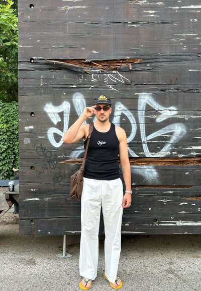 Top sin mangas negro, pantalones anchos blancos, bolsa de mensajero marrón, gafas de sol y una gorra negra. El fondo presenta madera texturizada con graffiti.
