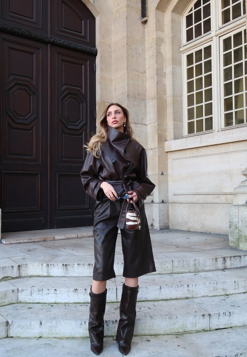 Femme debout sur des marches en pierre, portant un trench-coat en cuir marron foncé, un pantalon culotte assorti, des bottes montantes et tenant un petit sac à main à motif zébré.