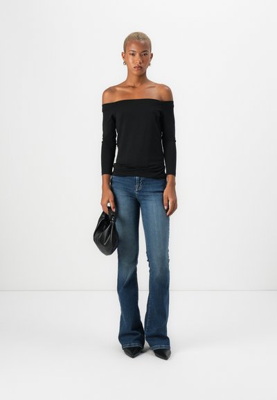 Top negro de manga larga y sin hombros combinado con jeans azules acampanados. La modelo sostiene un bolso negro; unos zapatos negros de punta completan el conjunto.