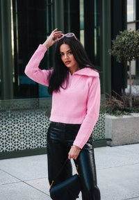 Rosa ribbet strikket sweater med lynlås krave, parret med sorte patentlæderbukser. Sort håndtaske, der holder solbriller, foran en moderne baggrund.