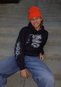 Melnā kapuce ar grafisku dizainu, kurā priekšpusē redzama silueta forma un zvaigžņu izlaušanās raksti uz piedurknes. Kombinēts ar gaiši ziliem džinsiem.