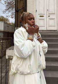 Hvid plisseret bomberjakke med høj krave, kombineret med en hvid plisseret kjole. Accessoires omfatter lagdelte armbånd og ringe. Grå trin i baggrunden.