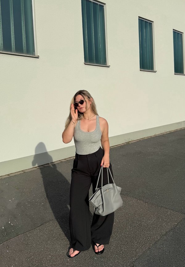 Femme portant des lunettes de soleil, un débardeur gris, un pantalon large noir et des tongs, tenant un grand sac à main gris, debout sur un trottoir près d'un bâtiment blanc.
