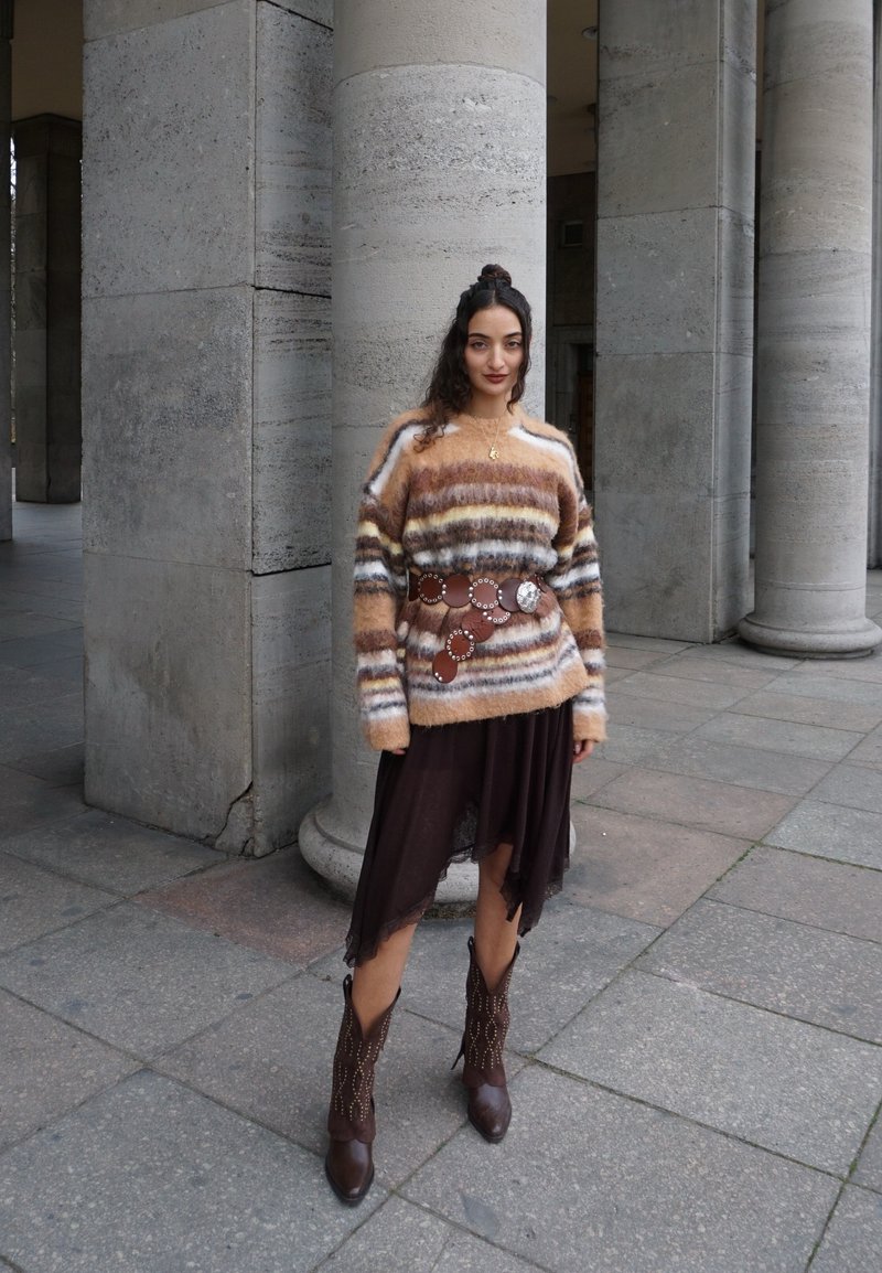 Vrouw met krullend haar in een gestreepte, pluizige trui, brede met studs versierde riem, asymmetrische donkere rok en bruine cowboylaarzen, staand bij stenen zuilen.