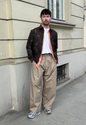 Hombre de pie en la acera de la ciudad con una chaqueta oscura estampada sobre una camisa durazno y una camiseta blanca, pantalones anchos beige y zapatillas grises.