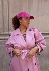 Trench coat rosa chiaro con vita legata, sovrapposto a un top a fantasia. Cappellino rosa con testo, gioielli dorati e una borsa viola. Capelli ricci.