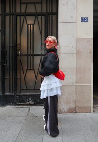 Personne aux cheveux roses courts portant des lunettes rouges, une doudoune noire, une jupe blanche à volants par-dessus un pantalon de survêtement noir, et tenant un sac rouge près d'une porte noire.