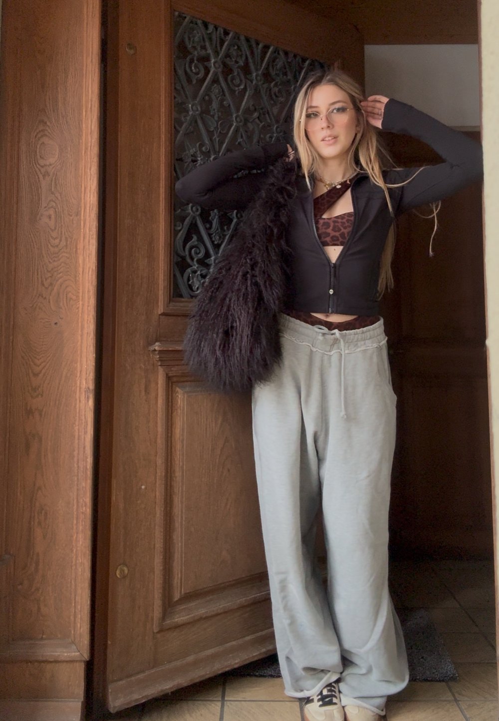 Jeune femme aux longs cheveux blonds portant des lunettes, un haut zippé noir, un bralette imprimé léopard, un pantalon de survêtement gris et des baskets blanches, debout près d'une porte en bois.