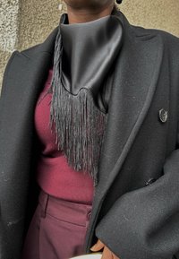 Personne portant un manteau noir, un haut et un pantalon bordeaux, avec une écharpe noire en soie ornée de longues franges autour du cou.