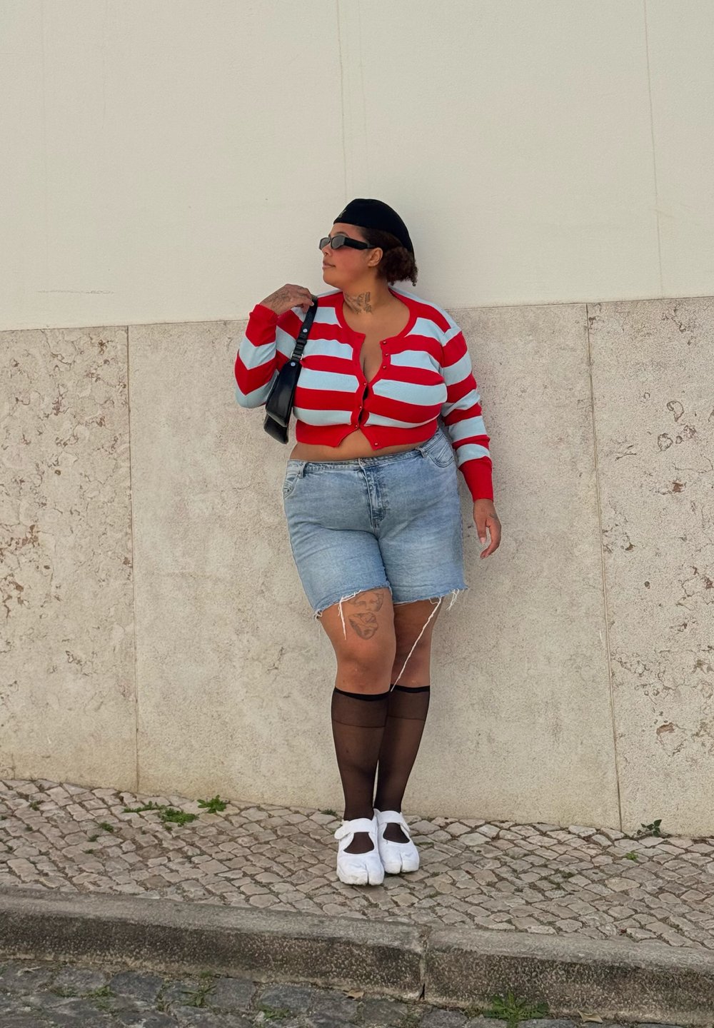 Cardigan court rayé rouge et bleu clair, short en denim, bas noirs jusqu'à mi-cuisse, chaussures blanches et petit sac noir.