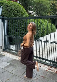 Brune, vide bukser parret med en brune mønstrede bluse med flæser. Lille grå håndtaske og slip-on sko med hvide striber.