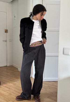 Chaqueta negra con bordado de flores, camiseta blanca, cinturilla estampada, pantalones grises holgados y zapatos marrones. Suelo de madera y paredes blancas en el fondo.