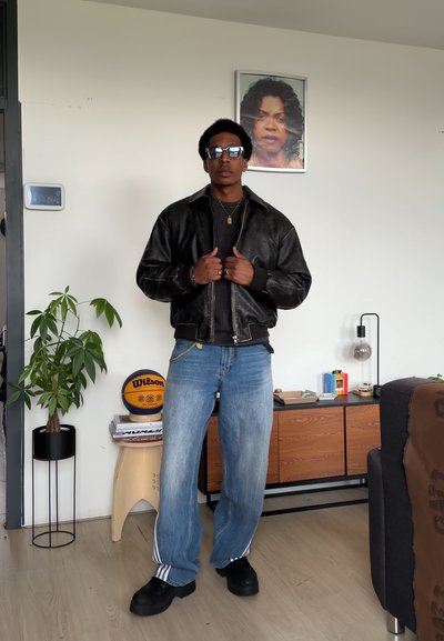 Chaqueta bomber de piel negra, camiseta negra de canalé, vaqueros holgados azul claro con rayas blancas, zapatos negros, collar plateado, entorno interior.