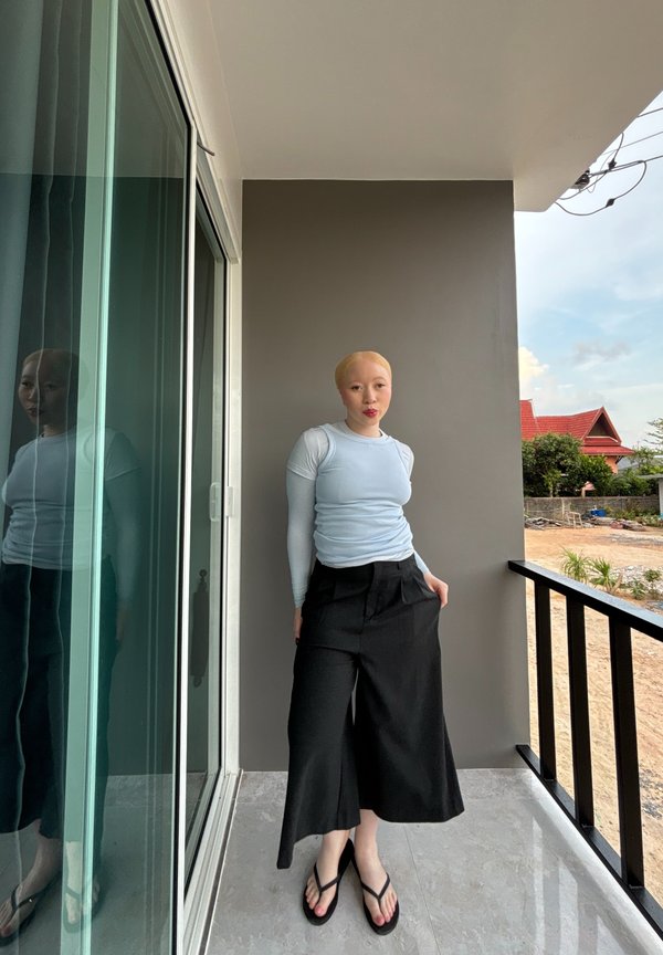 Person with short blonde hair wearing a light blue long-sleeved top and black wide-leg trousers stands on a balcony next to a sliding glass door.