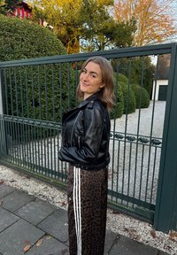 Chaqueta de cuero negra estilo motorista con cremalleras, combinada con una falda larga estampada de leopardo con rayas blancas. El fondo incluye vegetación y una puerta.