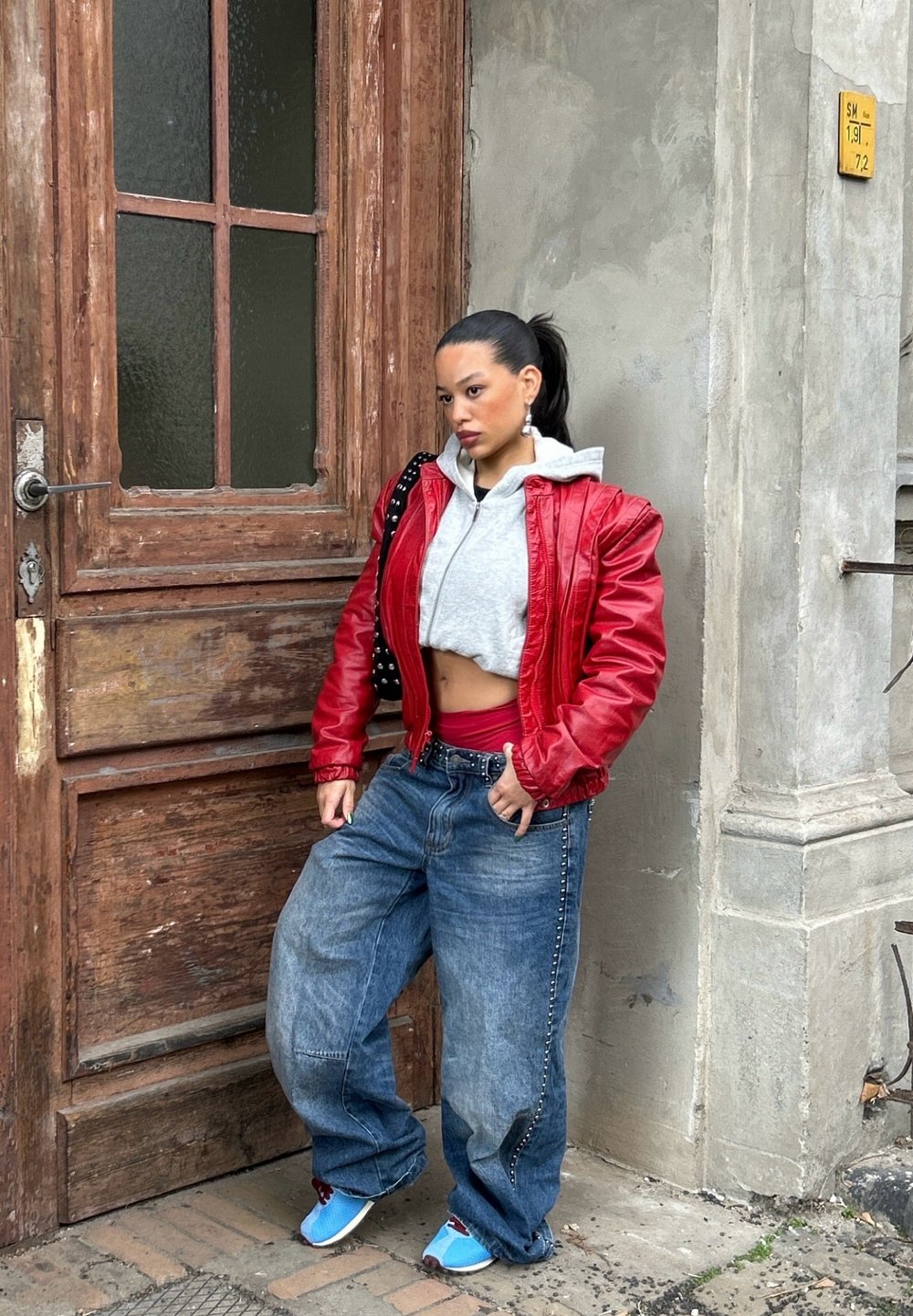 Giacca di pelle rossa sopra un hoodie grigio corto, abbinata a jeans blu oversize e scarpe blu. In piedi accanto a una porta di legno testurizzata.