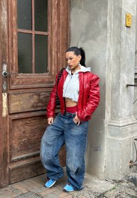 Giacca di pelle rossa sopra un hoodie grigio corto, abbinata a jeans blu oversize e scarpe blu. In piedi accanto a una porta di legno testurizzata.