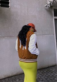 Ruskea ja valkoinen pommijakku, jossa on raidallinen helma, yhdistettynä tiukasti istuvaan keltaiseen hameeseen ja punaiseen lippikseen. Vihreät korvakorut tuovat asuun lisää ilmettä.