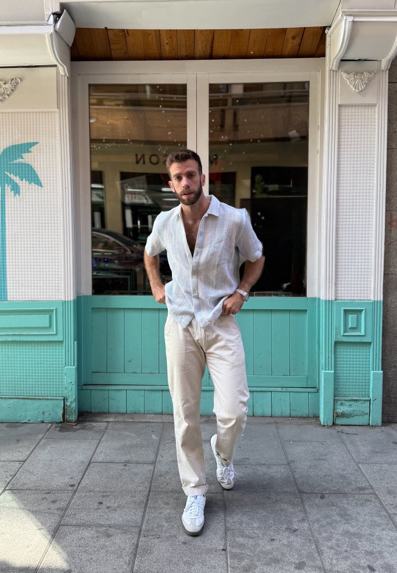 Camicia a maniche corte a scacchi azzurri, pantaloni crema e sneakers bianche. In piedi contro un muro turchese con dettagli in legno.
