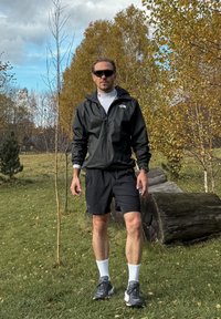 Black zip-up hooded jacket, white long-sleeve base layer, black shorts, white socks, and grey trainers, set against a natural outdoor background.