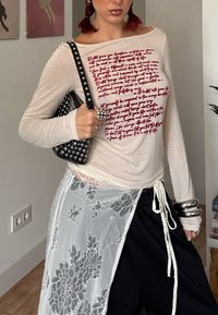 Haut beige à manches longues avec texte rouge, associé à un pantalon noir ample et un overlay en dentelle blanche transparente avec des motifs floraux. Sac à main noir clouté.