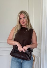 Hnědá kožená kabelka s řetízkovým detailem, obdélníkového tvaru, nošená s hnědou blůzkou s krátkým rukávem a světle modrými džíny na neutrálním pozadí.