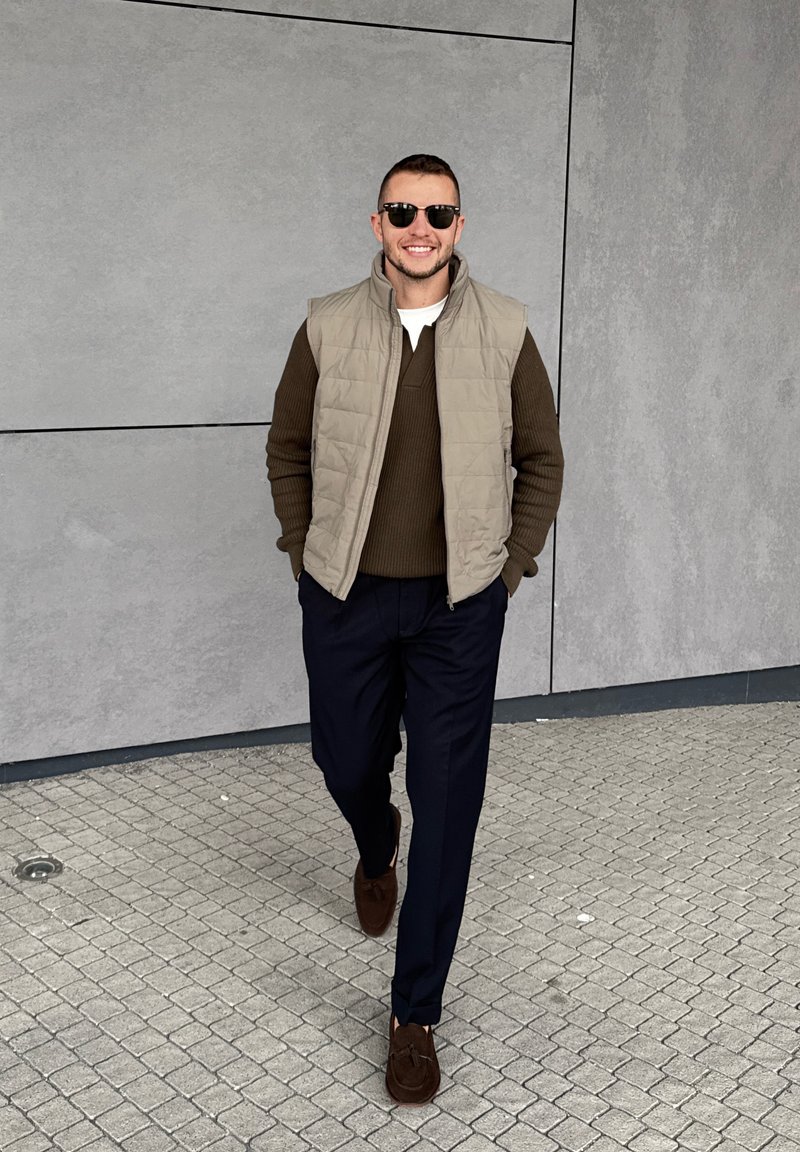 Man wearing sunglasses, beige waistcoat, brown jumper, navy trousers, and brown shoes walking on a tiled pavement by a grey wall.