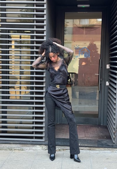 Top negro de malla de manga larga sobre un vestido negro de satén, combinado con pantalones de rayas finas y botas negras de tacón. Cuenta con un cinturón con detalle de logo.