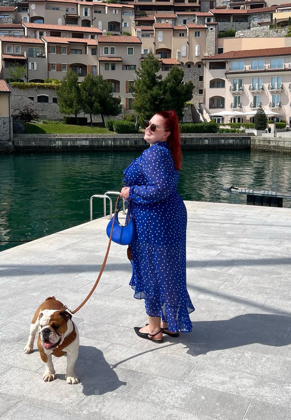Sininen pilkullinen mekko, jossa on läpinäkyvä kangas, frillatut helmat ja pitkät hihat; kantaa sinistä käsilaukkua; kävelee bulldogin kanssa hihnassa rannan läheisyydessä.