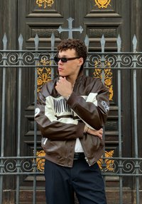 Hnedá kožená bomber bunda s bielou panelou, ktorá obsahuje text "MOTOR", rebrované manžety a pretekárske kockované detaily; nosená cez čierne nohavice.