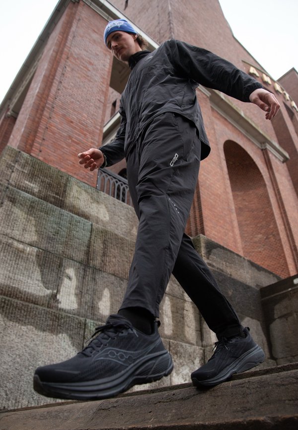Άνδρας με μαύρα αθλητικά ρούχα και αθλητικά παπούτσια περπατάει σε εξωτερικά σκαλοπάτια από πέτρα μπροστά από ένα κτίριο με τούβλα.