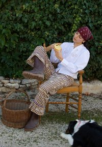Femme épluchant une pomme assise sur une chaise en bois en plein air, portant un bandana rouge, une chemise blanche, un pantalon à motif, avec un chien couché à proximité sur du gravier.