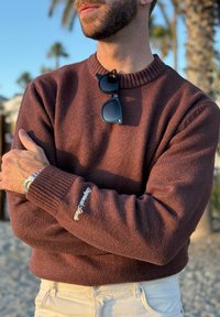 Homme barbu portant un pull bordeaux, un pantalon beige, une montre argentée, des lunettes de soleil noires accrochées au col, debout en plein air avec des palmiers en arrière-plan.